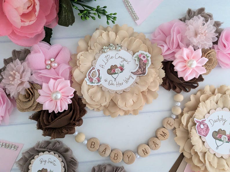 May include: A pink and brown baby shower decoration with a flower garland and a wooden bead garland that says "Baby Name". The flower garland has a brown flower with a white circle that says "A Little Cowgirl is On Her Way".