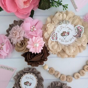 May include: A brown, pink, and beige flower crown with a pink and white "A Little Cowgirl is On Her Way" sign in the center.  The crown is decorated with rhinestones and pearls.  A wooden bead necklace with the words "BABY NAME" is also pictured.
