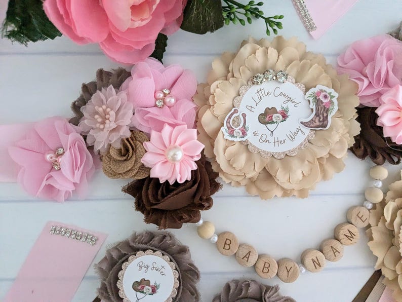 May include: A brown, pink, and beige flower crown with a pink and white "A Little Cowgirl is On Her Way" sign in the center.  The crown is decorated with rhinestones and pearls.  A wooden bead necklace with the words "BABY NAME" is also pictured.