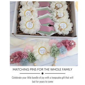 May include: A set of white fabric flower pins with gold accents and text that reads "Mommy to Be", "Daddy to Be", "Grandpa to Be", and "Big Sister". The pins are arranged in a box with pink and green ribbon. A white sash with a similar flower design and the text "Mommy to Be" is also included.
