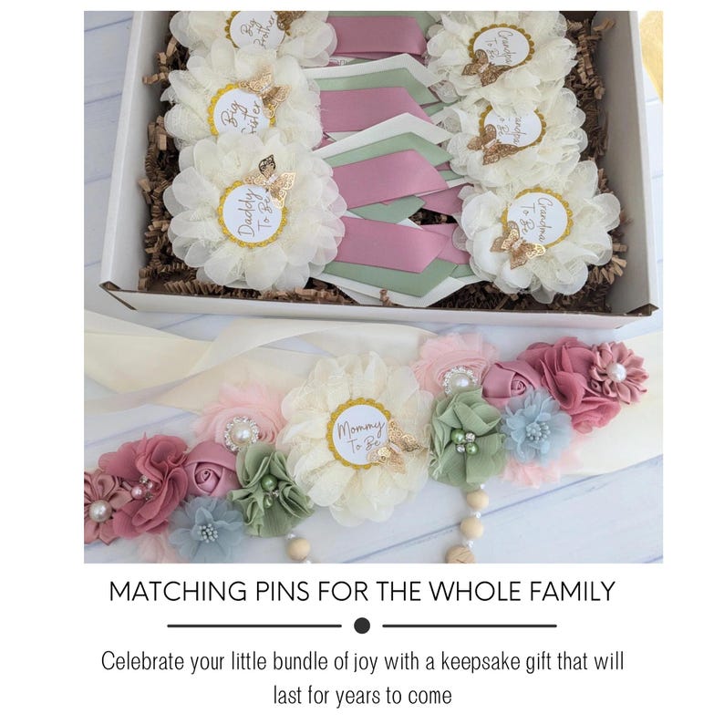 May include: A set of white fabric flower pins with gold accents and text that reads "Mommy to Be", "Daddy to Be", "Grandpa to Be", and "Big Sister". The pins are arranged in a box with pink and green ribbon. A white sash with a similar flower design and the text "Mommy to Be" is also included.