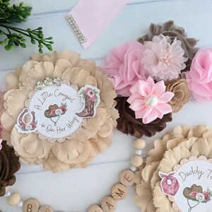 May include: A beige flower with a white circle in the center that says "A Little Cowgirl is On Her Way". There is a brown flower with pink flowers on top. The flowers are on a white surface with wooden letters that spell "NAME".