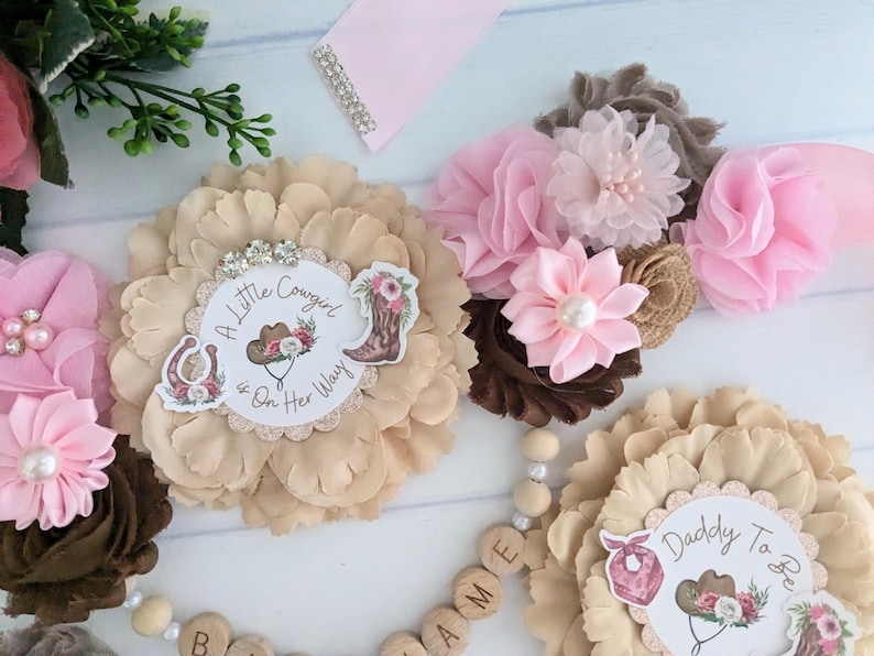 May include: A beige flower with a white circle in the center that says "A Little Cowgirl is On Her Way". There is a brown flower with pink flowers on top. The flowers are on a white surface with wooden letters that spell "NAME".