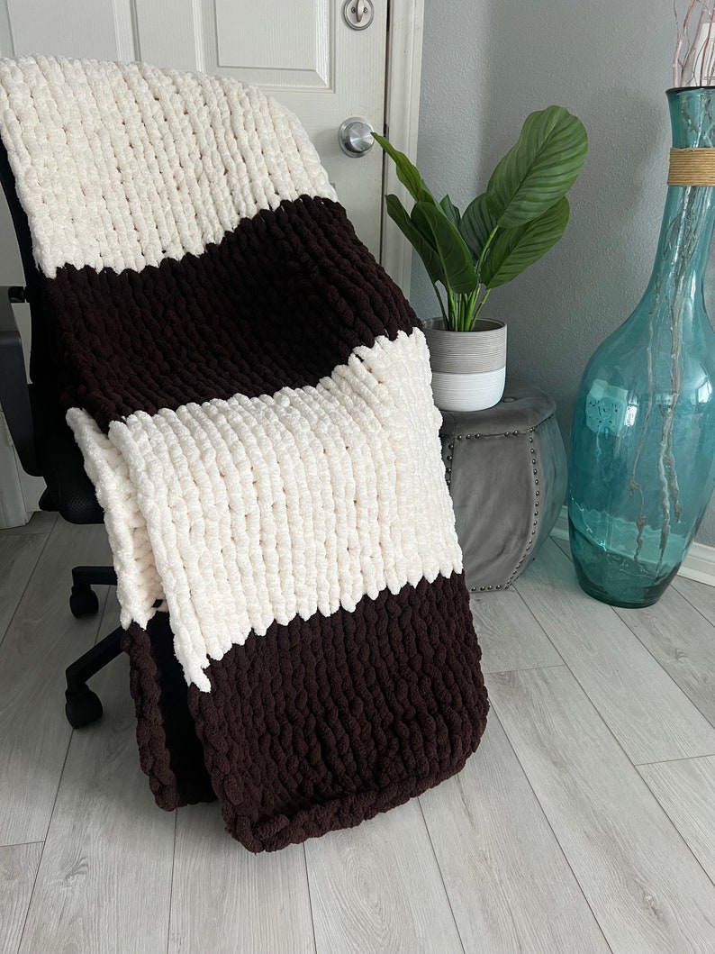 Może przedstawiać: Gruby, dzianinowy koc w stylu color block. Koc ma naprzemienne paski w kolorze kremowym i ciemnobrązowym. Koc jest narzucony na czarne krzesło, co podkreśla jego fakturę i kontrast kolorystyczny. Przytulny koc do dekoracji wnętrz.