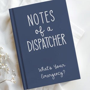 May include: A navy blue notebook with the text "NOTES of a DISPATCHER" in white script. Below, the notebook reads "What's Your Emergency?" The notebook is on a white surface with delicate white flowers in the corner.