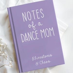 May include: A lavender notebook with the text "NOTES of a DANCE MOM" in white script. Below, the text "Rhinestones & Chaos" is also in white script. The notebook is on a white surface with small white flowers.