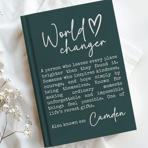 May include: A dark teal journal with the words "World Changer" and a heart symbol in white script. The journal includes a definition of a world changer and the name "Camden" in white script. The journal is on a white cloth.