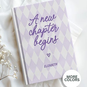 May include: A hardcover book with a lavender and white harlequin pattern. The cover features the text "A new chapter begins" in purple script, a small heart, and the name "Elizabeth".
