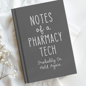 May include: A gray notebook with the text "NOTES of a PHARMACY TECH Probably On Hold Again" in white. The notebook is on a white surface with small white flowers.