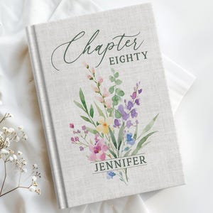 May include: A hardcover book with a light gray linen-textured cover featuring a watercolor floral bouquet in shades of pink, purple, yellow, and green. The text reads Chapter EIGHTY and the name JENNIFER in dark gray serif and script fonts.