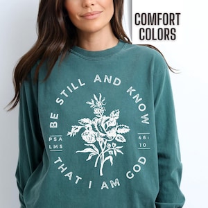 May include: Teal long-sleeve shirt with a white floral design and the text "Be Still and Know That I Am God" with a Bible verse reference.