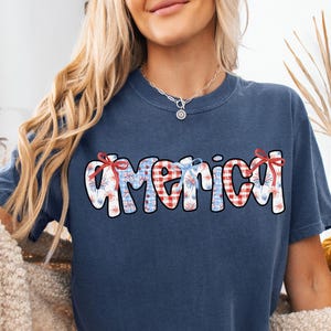 May include: A navy blue t-shirt with a red, white, and blue "America" graphic featuring a checkered and floral pattern.