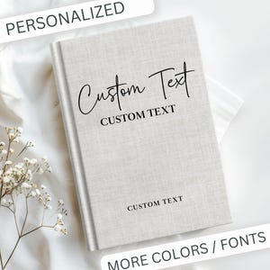 May include: A light gray hardcover journal with the words "Custom Text" in a script font. The journal has the words "Custom Text" printed on the cover and spine. The image also includes the words "Personalized" and "More Colors / Fonts".