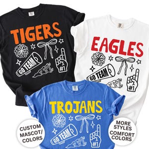 Personalized school spirit shirt displayed on a Comfort Colors garment featuring a custom school mascot name surrounded by colorful school spirit doodles. Buyers can personalize the mascot text and choose colors to match their school's team colors. Available as an adult Comfort Colors t-shirt, long sleeve tee, sweatshirt, and youth short sleeve tee. Perfect for football games, basketball season, pep rallies, spirit week, school events, and showing school pride all year long.