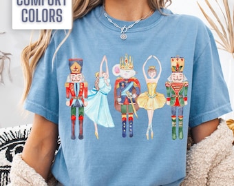 May include: A light blue t-shirt with a watercolor illustration of a ballerina, a nutcracker, and a mouse king. The text "Comfort Colors" is printed on a white tag.