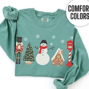 May include: Light green long-sleeve t-shirt with a Christmas-themed design featuring a snowman, a gingerbread house, a nutcracker, and a Christmas tree. The shirt has the text "COMFORT COLORS" printed on the top right corner.