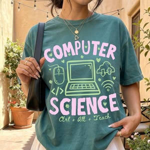 May include: A dark green t-shirt featuring a light green graphic of a laptop, a computer mouse, and cloud icons. The text reads "COMPUTER SCIENCE" in large pink letters, with "Ctrl + Alt + Teach" in smaller green text below the laptop graphic.