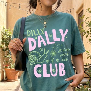 Colorful Dilly Dally Club graphic shirt featuring bright playful lettering and fun whimsical artwork printed on Comfort Colors apparel. Trendy graphic tee available as a short sleeve t-shirt, long sleeve shirt, sweatshirt, and youth tee. Fun casual design with colorful details perfect for everyday wear, summer outfits, relaxed weekends, school, vacations, and gifting. Cute preppy style shirt for women, teens, girls, moms, and anyone who loves cheerful, humorous graphic apparel.