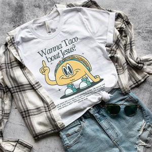 May include: White t-shirt with a cartoon taco graphic and the text "Wanna Taco bout Jesus?"  The taco is wearing sneakers and has a surprised expression. The text "For God so loved the world that He gave His one and only Son, that whoever believes in Him shall not perish but have eternal life. John 3:16" is printed below the graphic.