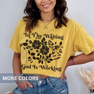 Op de afbeelding: Een mosterdgeel T-shirt met een zwart bloemenontwerp en de tekst "In the waiting God is working".