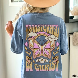 May include: A blue jean t-shirt with a graphic of a butterfly, rainbow, and clouds. The text "TRANSFORMED BY CHRIST" is written in a retro font. The text "ROMANS 12:2" is written in a smaller font.