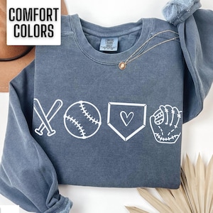 May include: A dark blue sweatshirt with a white baseball-themed graphic. The graphic includes a baseball, bats, a baseball glove, and a heart inside a home plate. The text "COMFORT COLORS" is printed on a white tag.