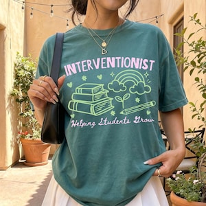 May include: A dark green t-shirt featuring the text INTERVENTIONIST in pink and Helping Students Grow in white script. The graphic includes a stack of three books, a rainbow, and a pencil in light green, accented with small hearts and stars.