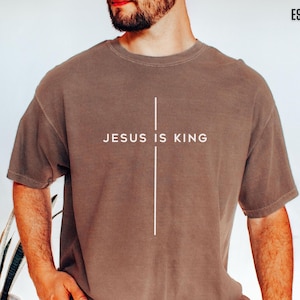 Op de afbeelding: Espresso-kleurig t-shirt met de witte tekst "JESUS IS KING" gecentreerd op de borst. Een verticale witte lijn kruist de tekst. Het shirt heeft korte mouwen en een ronde hals.
