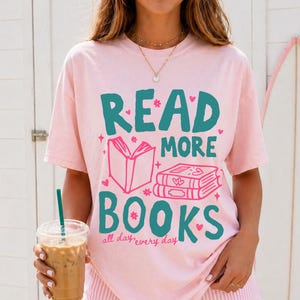 May include: A light pink short-sleeve t-shirt featuring the teal and pink text "READ MORE BOOKS" along with illustrations of an open book and a stack of books. The shirt also includes the small pink text "all day, every day" below the main graphic.