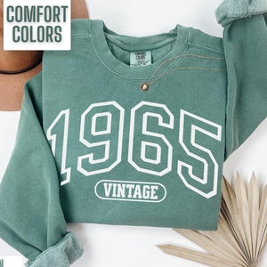 May include: A green sweatshirt with the year 1965 printed in white on the front. The sweatshirt also has the word "Vintage" printed in white on a small rectangle.