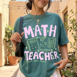 May include: A dark green short-sleeve t-shirt featuring the pink text MATH TEACHER with graphic illustrations of a notebook, a calculator, a pencil, and the numbers 1, 2, and 3. The shirt also includes the cursive text Teach, Love, Repeat.