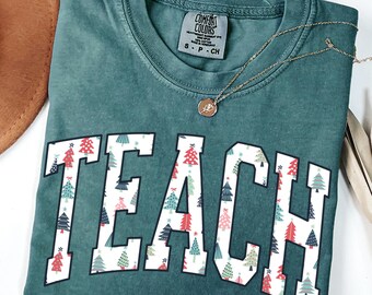 May include: Blue spruce t-shirt with the word "TEACH" in white block letters filled with Christmas tree patterns. A gold necklace with a pendant rests on the shirt. The shirt is folded on a white surface.