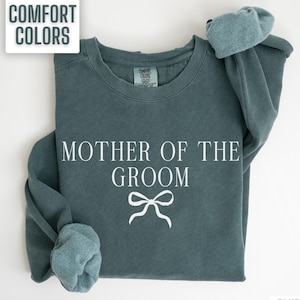 May include: A teal blue sweatshirt with the text "Mother of the Groom" in white lettering and a white bow. The sweatshirt is made of a soft, comfortable material.