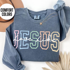 May include: A dark blue sweatshirt featuring the word "JESUS" in rainbow colours and the words "love like" written in white cursive underneath. The sweatshirt is made from a soft, comfortable fabric and has a relaxed fit.
