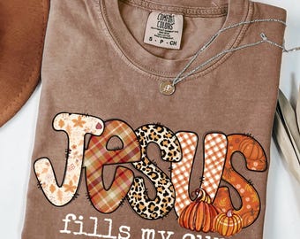 May include: Espresso-colored t-shirt with the words "JESUS fills my cup" in a decorative font. The word "JESUS" features a mix of patterns, including fall leaves, plaid, and leopard print. Two pumpkins are below the word "JESUS". A silver necklace is on the shirt.