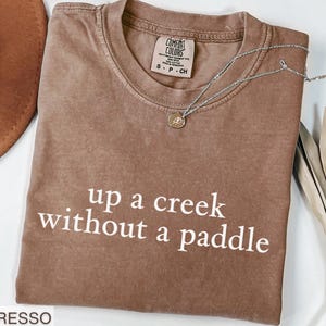 This funny shirt says up a creek without a paddle, just like the southern saying that is said in the south. This would be a funny gag gift for a friend, mom, sister or coworker.  This Comfort Colors shirt comes in 6 beautiful colors, espresso, light green, moss, blue jean, violet and pepper.  This humor tee will make a humorous gift. Share your sarcasm through your sarcastic apparel.