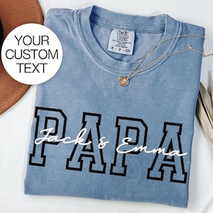 Op de afbeelding: Een lichtblauw t-shirt met het woord "PAPA" in zwarte blokletters en de namen "Jack & Emma" in witte letters. Een gouden ketting en een label zijn zichtbaar. Het shirt is opgevouwen en op een wit oppervlak geplaatst.