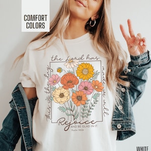May include: White t-shirt with a floral design and the text "This is the day the Lord has made we will rejoice and be glad in it. Psalm 118:24"