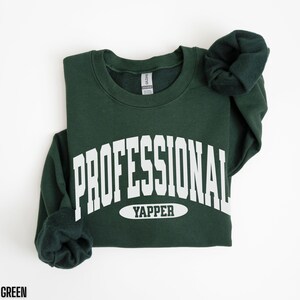 May include: Forest green sweatshirt with the text "PROFESSIONAL YAPPER" in white block letters. The sweatshirt is folded, with the sleeves bunched up. The text "FOREST GREEN" is at the bottom.
