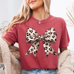 Puede incluir: Una camiseta roja con un gran lazo de estampado de leopardo marr&oacute;n y blanco en la parte delantera.