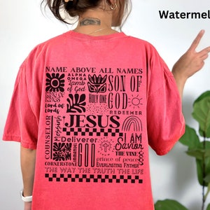 May include: A red t-shirt with a black graphic print of the word "JESUS" surrounded by other religious phrases and symbols. The text includes "Name Above All Names", "Son of God", "Holy One", "Redeemer", "Counselor", "Deliverer", "I Am Savior", "The Vine", "Prince of Peace", "Everlasting Father", "The Way The Truth The Life".
