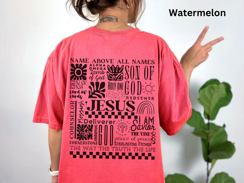 May include: A red t-shirt with a black graphic print of the word "JESUS" surrounded by other religious phrases and symbols. The text includes "Name Above All Names", "Son of God", "Holy One", "Redeemer", "Counselor", "Deliverer", "I Am Savior", "The Vine", "Prince of Peace", "Everlasting Father", "The Way The Truth The Life".