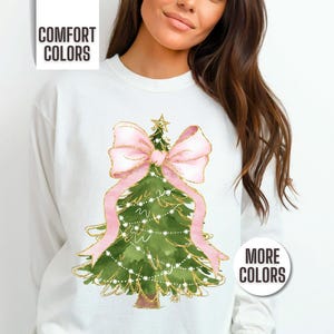 K&ouml;nnte beinhalten: Wei&szlig;es Sweatshirt mit einem Aquarell-Weihnachtsbaum-Design. Der Baum ist gr&uuml;n mit goldenen Akzenten, einer rosa Schleife und Lichterketten. Der Text "COMFORT COLORS" und "MORE COLORS" ist ebenfalls sichtbar.