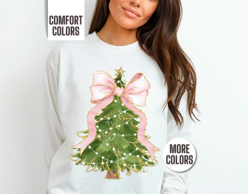 K&ouml;nnte beinhalten: Wei&szlig;es Sweatshirt mit einem Aquarell-Weihnachtsbaum-Design. Der Baum ist gr&uuml;n mit goldenen Akzenten, einer rosa Schleife und Lichterketten. Der Text "COMFORT COLORS" und "MORE COLORS" ist ebenfalls sichtbar.