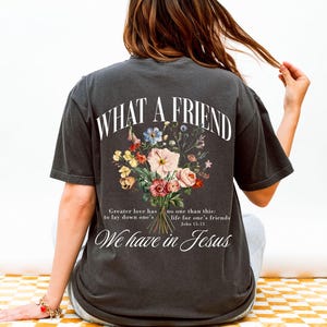 K&ouml;nnte beinhalten: Dunkelgraues T-Shirt mit dem Text "WHAT A FRIEND" &uuml;ber einem Blumenstrau&szlig;-Design. Darunter steht "We have in Jesus" und ein Bibelvers. Das Shirt hat einen l&auml;ssigen Vintage-Stil.