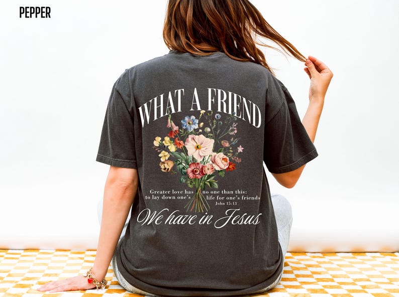 K&ouml;nnte beinhalten: Dunkelgraues T-Shirt mit dem Text "WHAT A FRIEND" &uuml;ber einem Blumenstrau&szlig;-Design. Darunter steht "We have in Jesus" und ein Bibelvers. Das Shirt hat einen l&auml;ssigen Vintage-Stil.