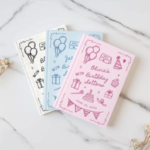 May include: Three personalized birthday journals in white, light blue, and pink. Each journal features hand-drawn illustrations of balloons, gifts, and a cake. The pink journal is labeled "Olivia's Birthday Letters" with a date of June 25, 2025.