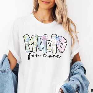 Op de afbeelding: Wit T-shirt met de tekst "MADE for more" in een kleurrijk ontwerp met bloemen- en streepmotieven. Het T-shirt heeft een relaxte pasvorm.