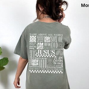 May include: A green t-shirt with a white graphic print of the word "JESUS" surrounded by other words and phrases, including "Name Above All Names", "Son of God", "Holy One", "Redeemer", "Deliverer", "I Am The Savior", "The Vine", "Prince of Peace", "Everlasting Father", "The Way The Truth The Life", and "Counselor, Messiah". The shirt is a relaxed fit and has a crew neck.