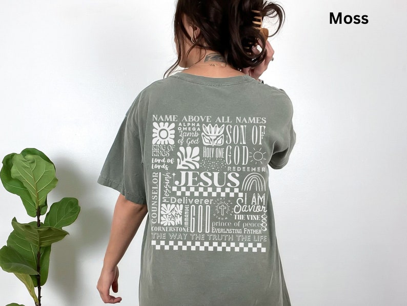 May include: A green t-shirt with a white graphic print of the word "JESUS" surrounded by other words and phrases, including "Name Above All Names", "Son of God", "Holy One", "Redeemer", "Deliverer", "I Am The Savior", "The Vine", "Prince of Peace", "Everlasting Father", "The Way The Truth The Life", and "Counselor, Messiah". The shirt is a relaxed fit and has a crew neck.